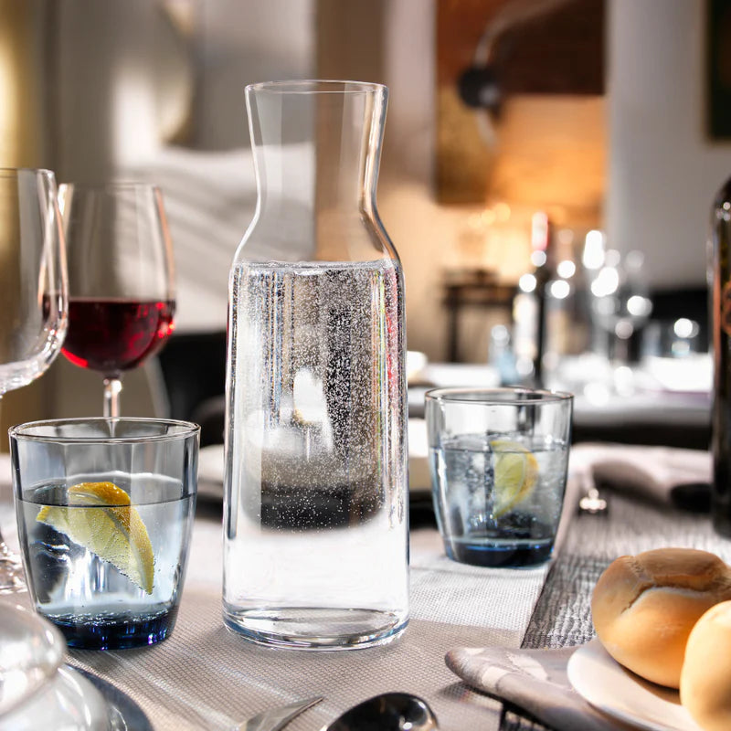 Clear glass carafe with water and lemon slices on a dining table with wine glasses and bread.