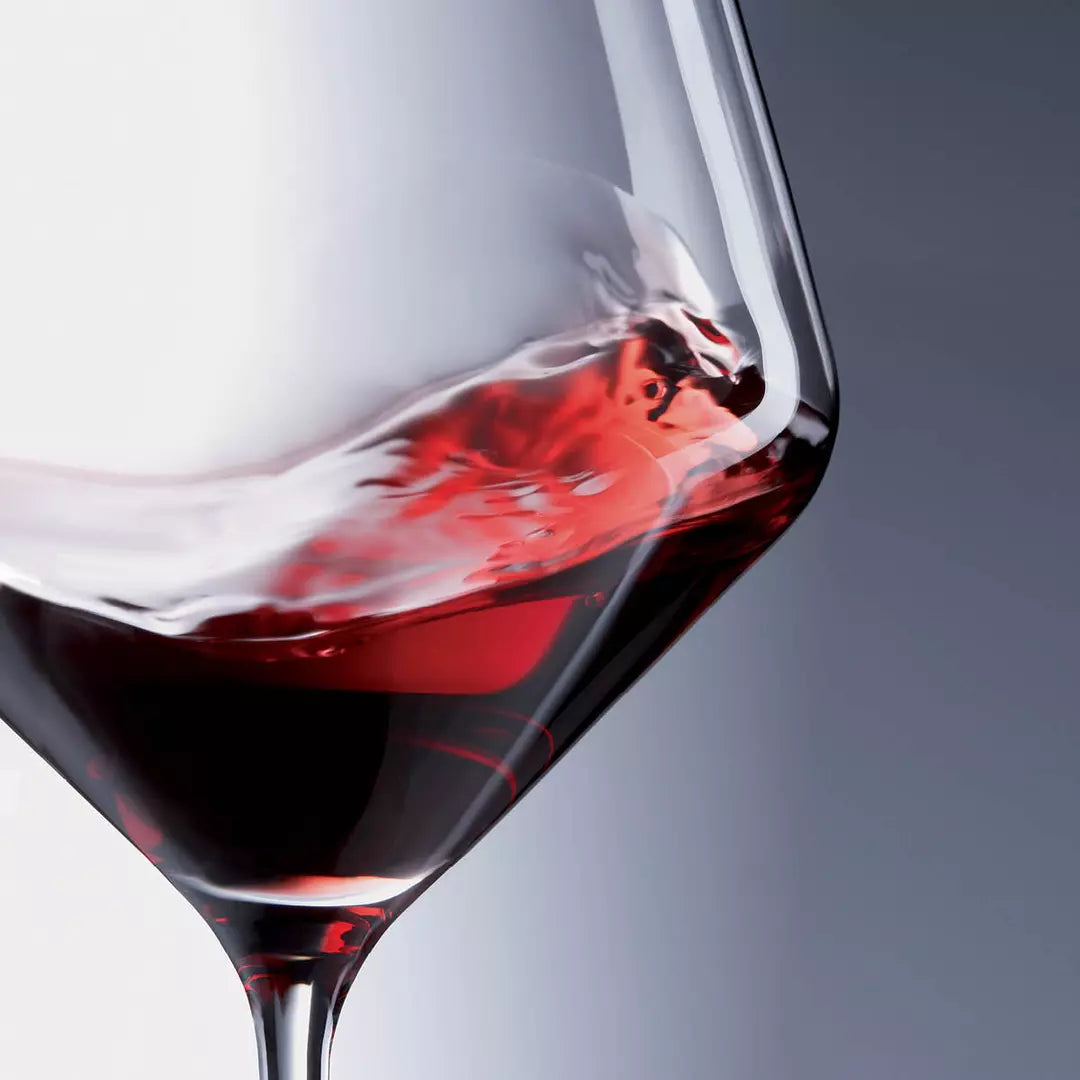 A close-up studio shot of a clear wine glass containing deep red wine, with the liquid swirling dynamically inside the bowl against a soft gray gradient background.