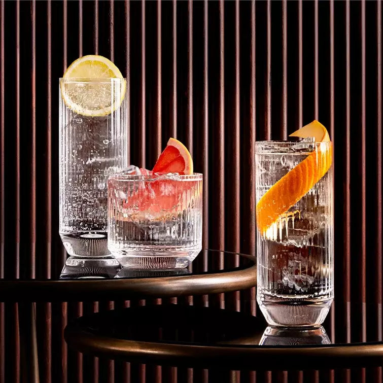 Three glasses with garnishes on a dark surface with vertical blinds in the background