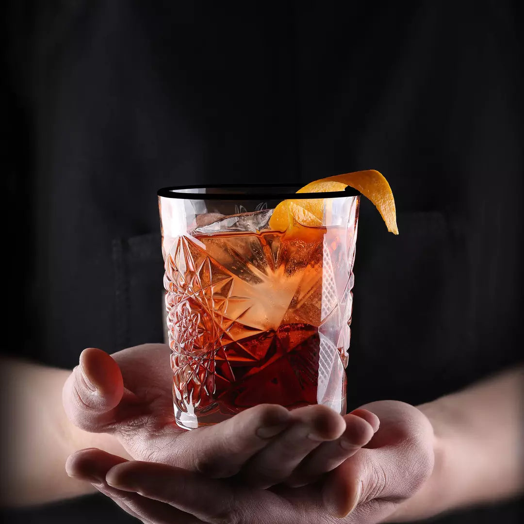 A hand holds a clear cut-glass tumbler containing an amber-brown cocktail with a large ice cube and an orange peel garnish, against a dark black background.