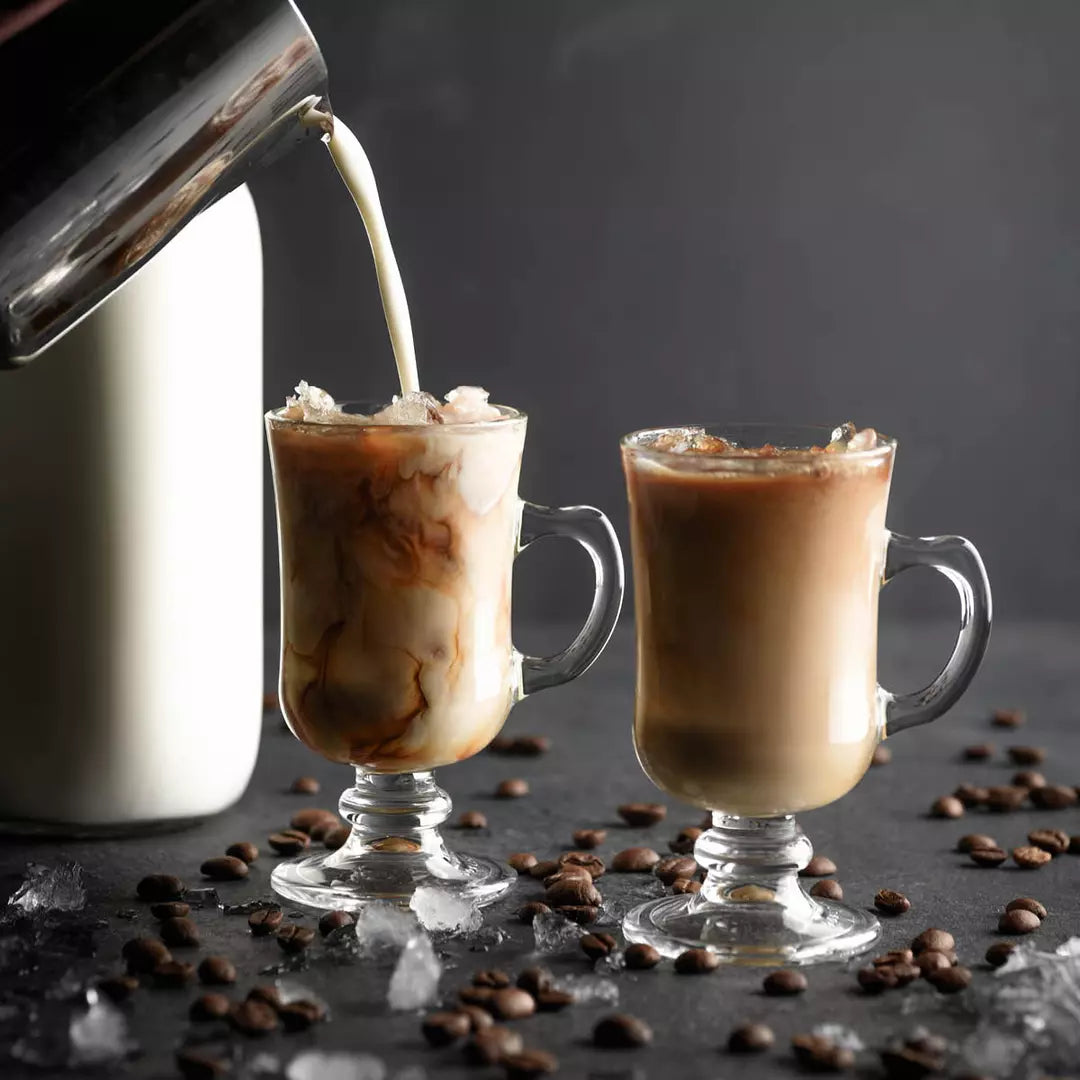 Iced coffee being poured into two glasses with ice and coffee beans on a dark surface.