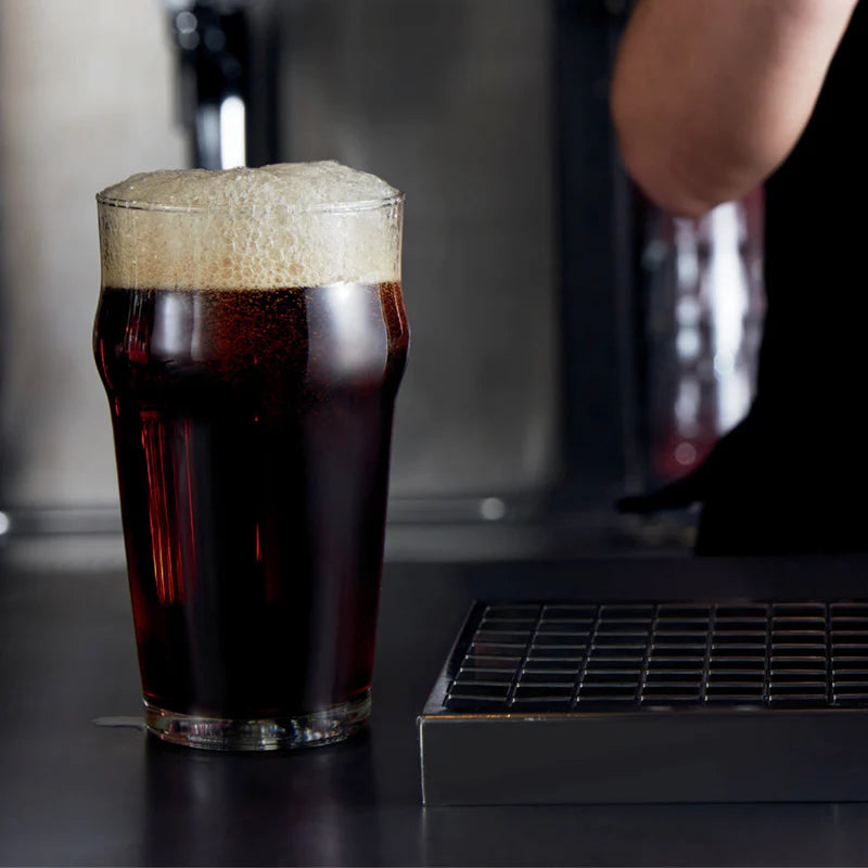 A pint glass filled with dark beer and topped with thick foam, set on a bar counter with a metal drain and a blurred bartender's arm in the background.