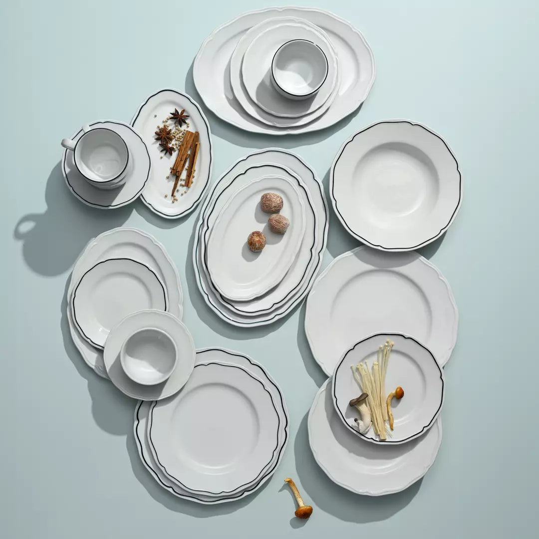 A flat-lay arrangement of a white ceramic dinnerware set with scalloped edges and thin dark rims, including plates, bowls, and cups, styled with small food props on a pale blue background.