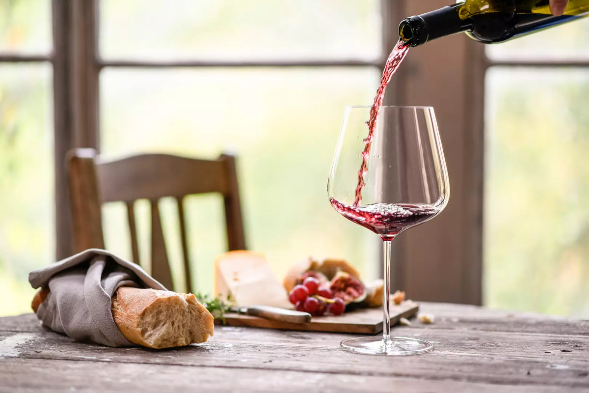 Rural table setting of a loaf of bread and cheese plate with a splash of red wine being poured into a large glass