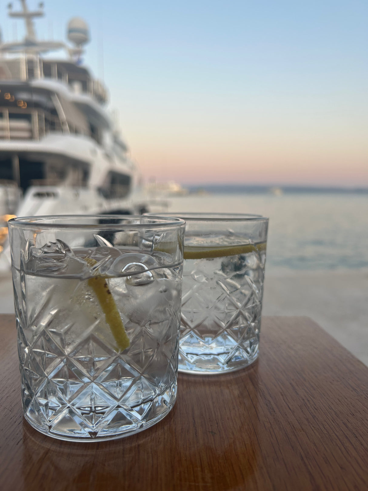 Two glasses with lemon slices on a table by a body of water with a boat in the background.