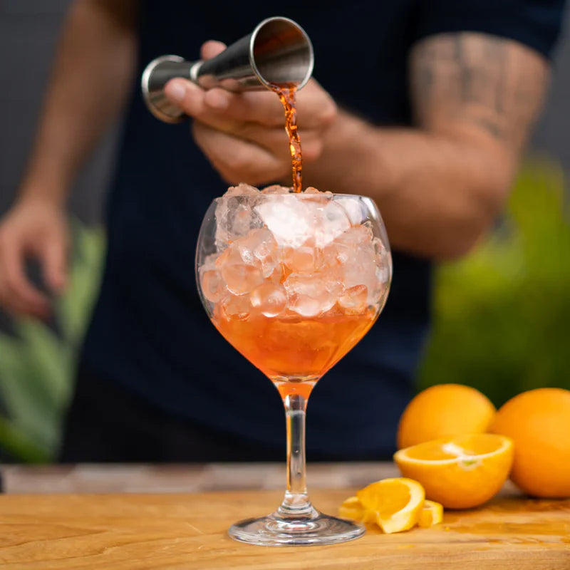 Cocktail being poured into a glass with ice, surrounded by oranges on a wooden surface.