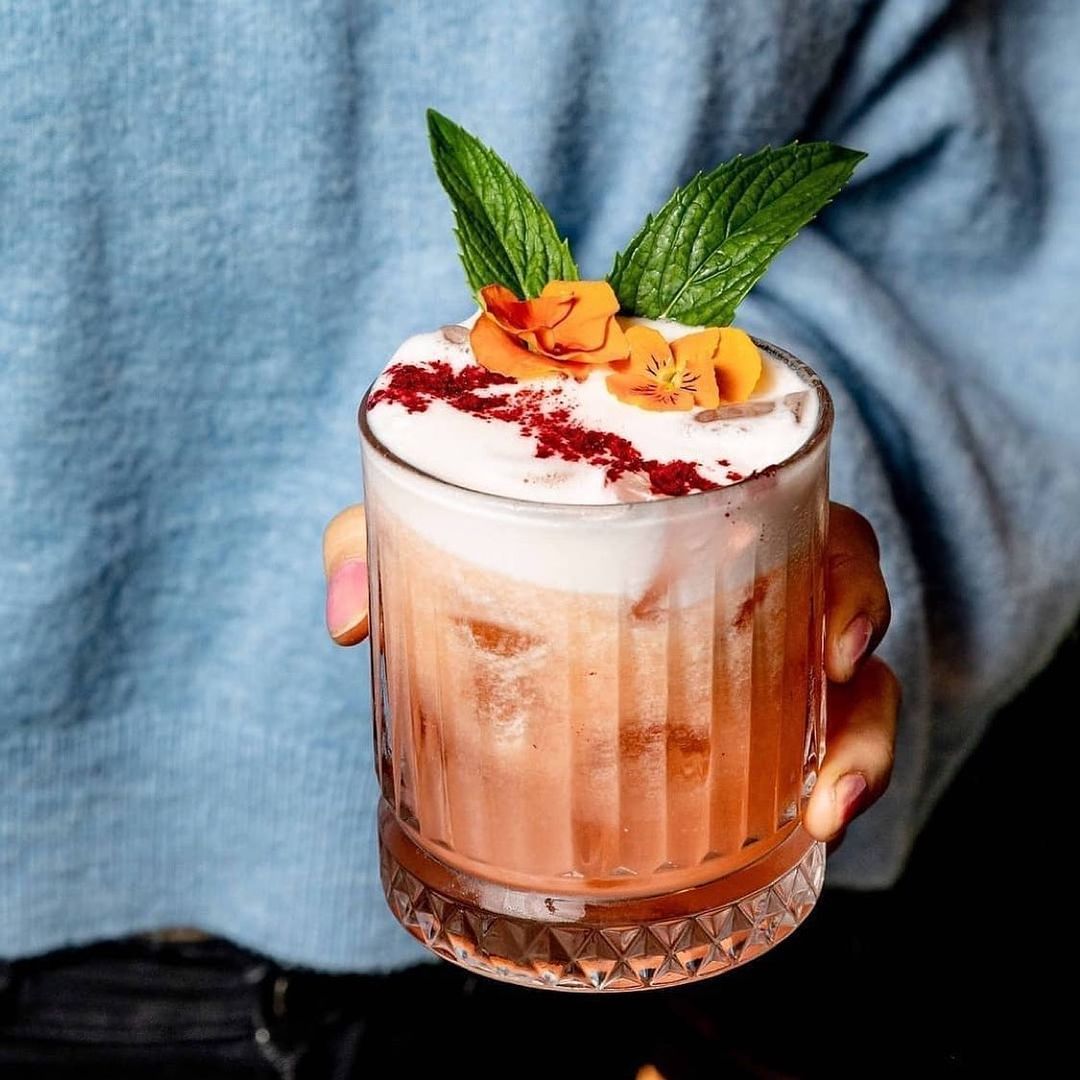 Hand holding a Pasabahce Elysia whisky glass containing a foamy pink cocktail topped with red berry dust, edible flowers and fresh mint leaves"