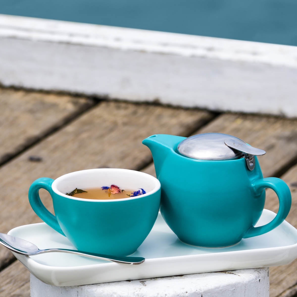 A medium sized aqua coloured coffee cup with liquid inside matched with the small coloured teapot on a white presentation tray