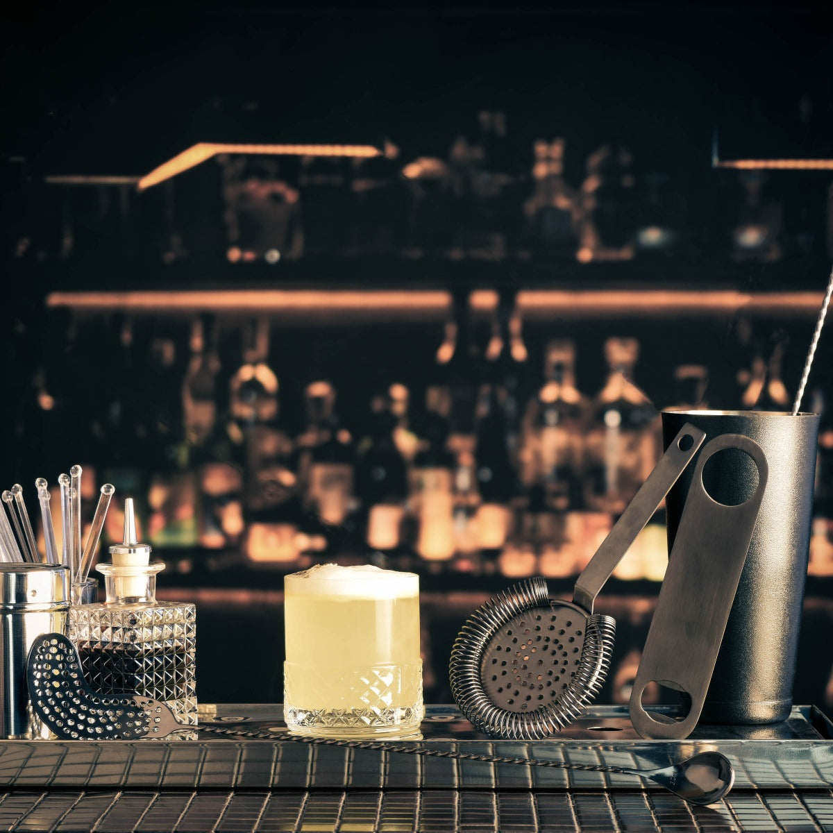 A range of cocktail making accessories on a bar counter with faded bottles and glasses in the background