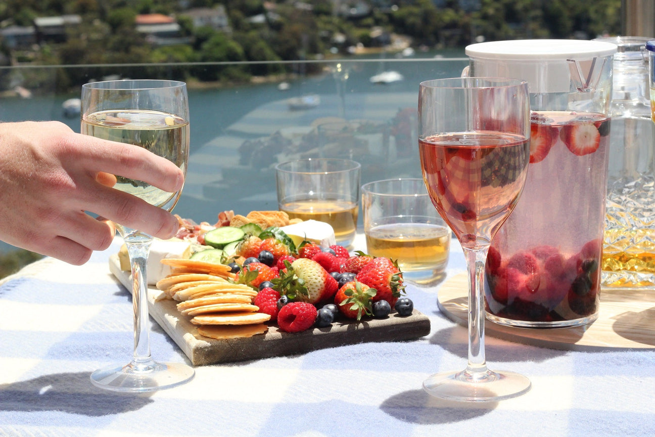 A picnic layout with cheese board, filled wine glasses and jugs with harbour background
