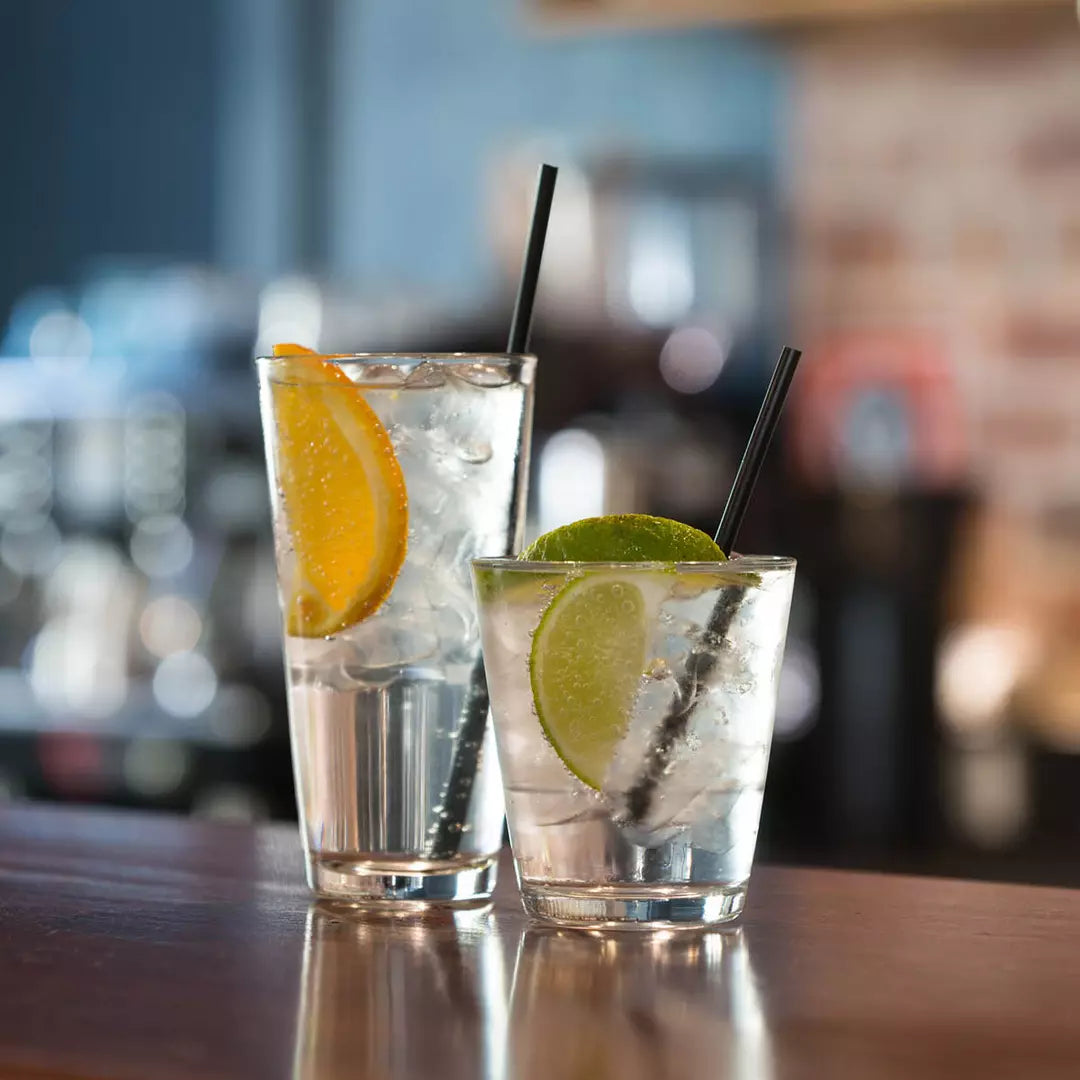 One large and one small tapered tumblers both with liquid, garnish and straws presented on a blurred bar setting