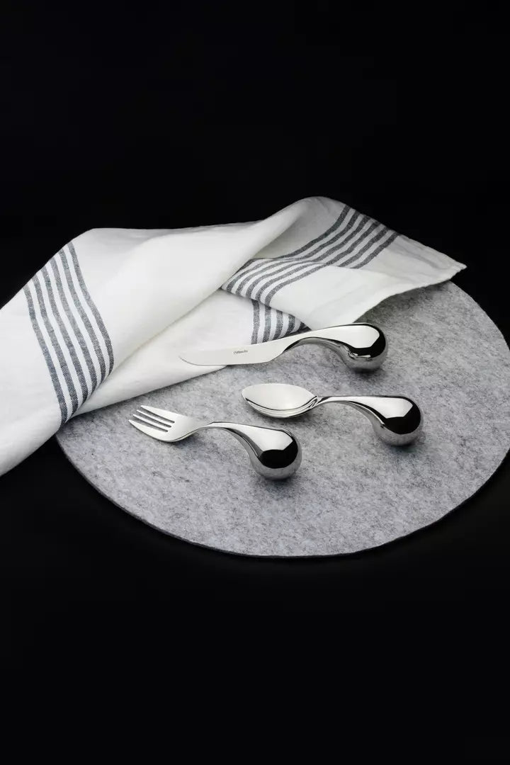 Three polished metallic utensils, including a fork, spoon, and a small knife, arranged on a round gray felt placemat with a white cloth napkin featuring gray stripes, set against a solid black background.