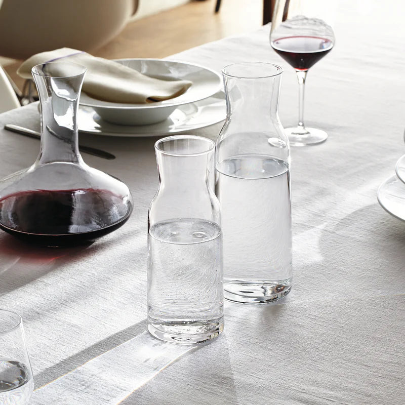 Clear glass carafes and decanter on a dining table with a bowl and wine glass in the background.