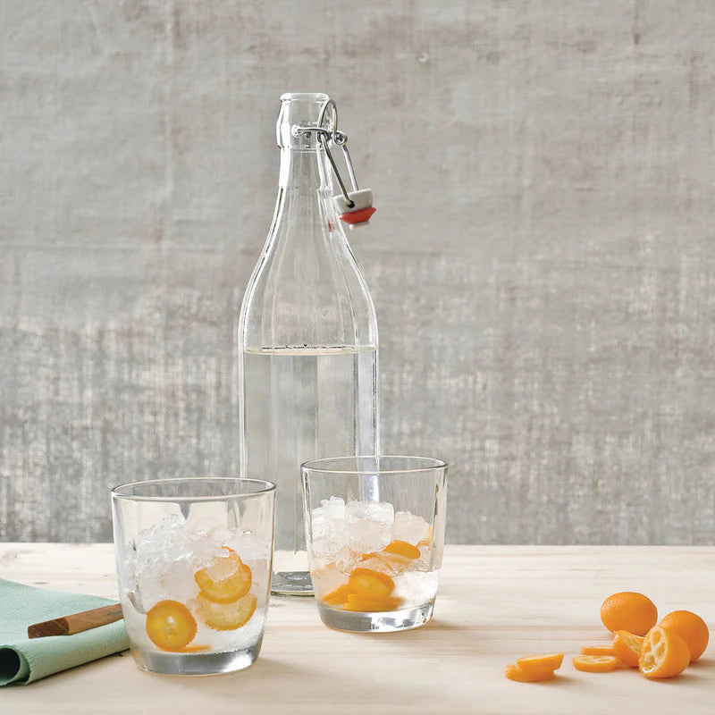 Clear glass bottle with a swing-top lid on a wooden surface with two glasses of iced water and orange slices.
