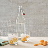 Clear glass bottle with a swing-top lid on a wooden surface with two glasses of iced water and orange slices.