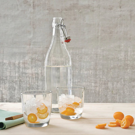 Clear glass bottle with a swing-top lid on a wooden surface with two glasses of iced water and orange slices.