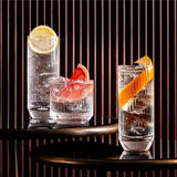 Three glasses with garnishes on a dark surface with vertical blinds in the background