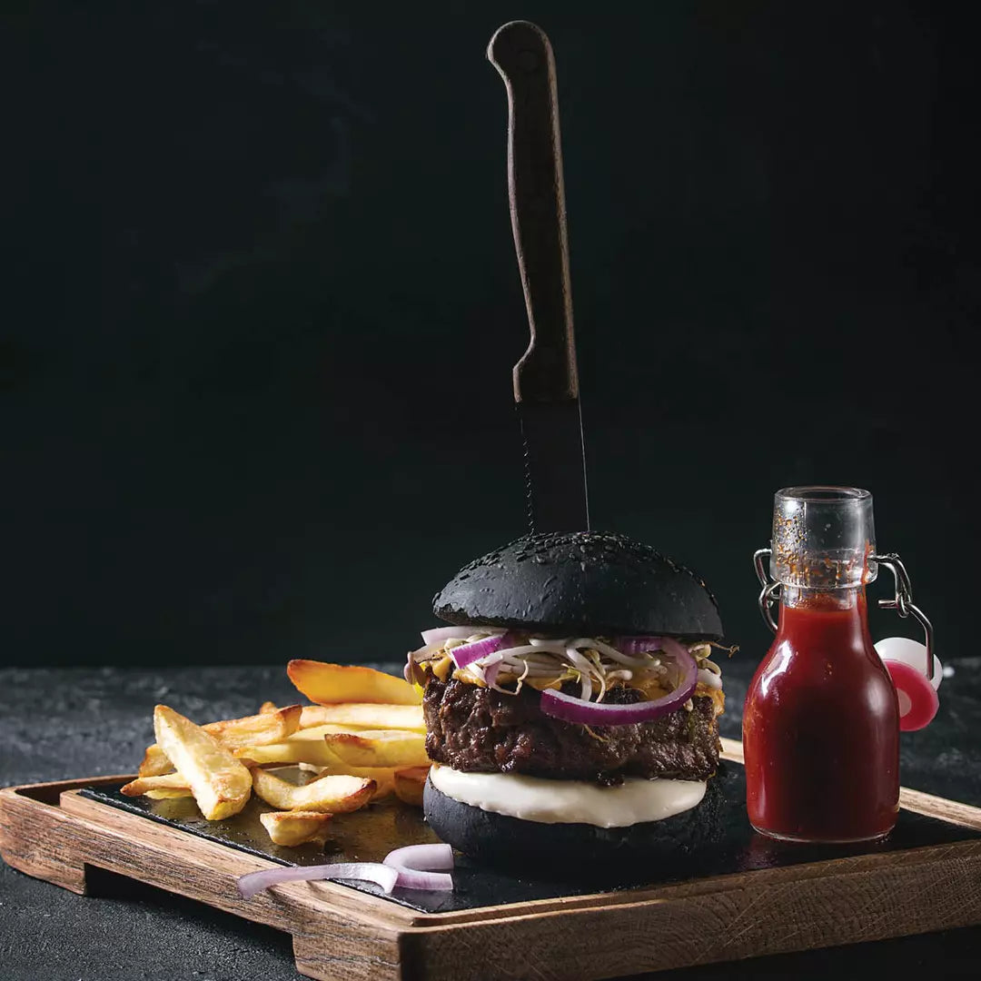Black burger with fries and a bottle of ketchup on a wooden board against a dark background