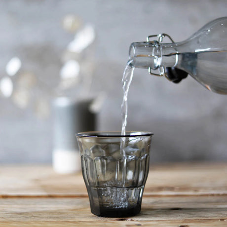 A clear tapered tumbler being fill with water from a bottle on a blurred background