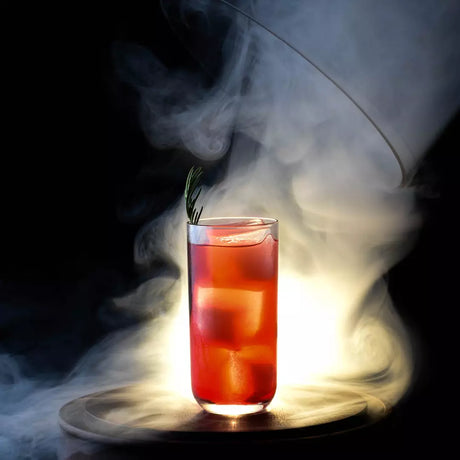A tall clear glass filled with a red-orange drink and large ice cubes, garnished with a sprig of rosemary, set on a dark surface with dramatic smoke rising around it against a black background.