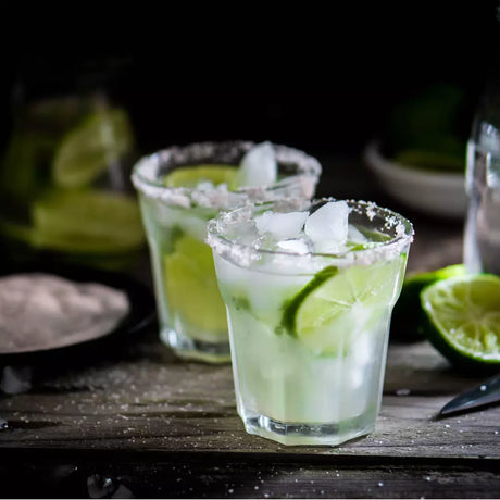 Two glasses of lime cocktails with ice and salt rims on a wooden surface.