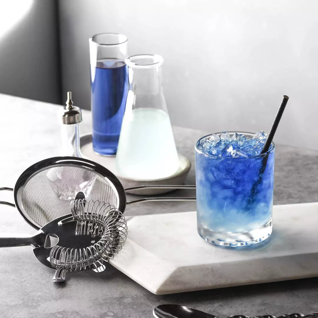 Bartending setup with a glass of blue cocktail, ice cubes, and bar tools on a marble surface.