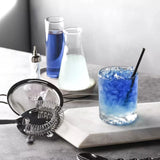 Bartending setup with a glass of blue cocktail, ice cubes, and bar tools on a marble surface.