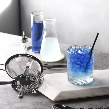 Bartending setup with a glass of blue cocktail, ice cubes, and bar tools on a marble surface.