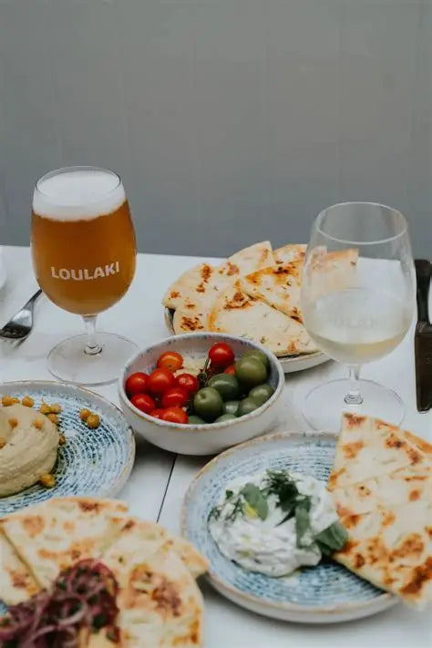 Table with Greek food and drinks including pita, hummus, and a glass of Loulaki beer.