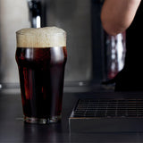 A pint glass filled with dark beer and topped with thick foam, set on a bar counter with a metal drain and a blurred bartender's arm in the background.