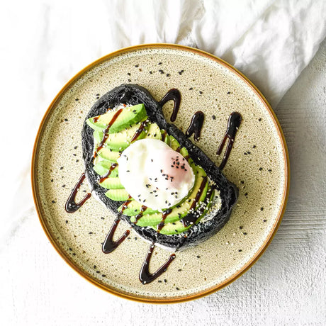 Top view of a medium sized brown speckled plate with Avocado and an egg on black bread on a white background