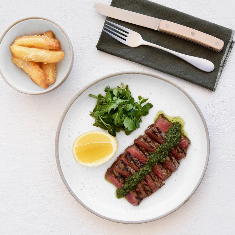 Steak with green sauce, lemon wedge, and salad on a plate with fries in the background.