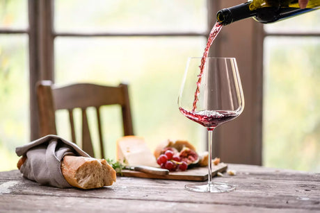 Rural table setting of a loaf of bread and cheese plate with a splash of red wine being poured into a large glass