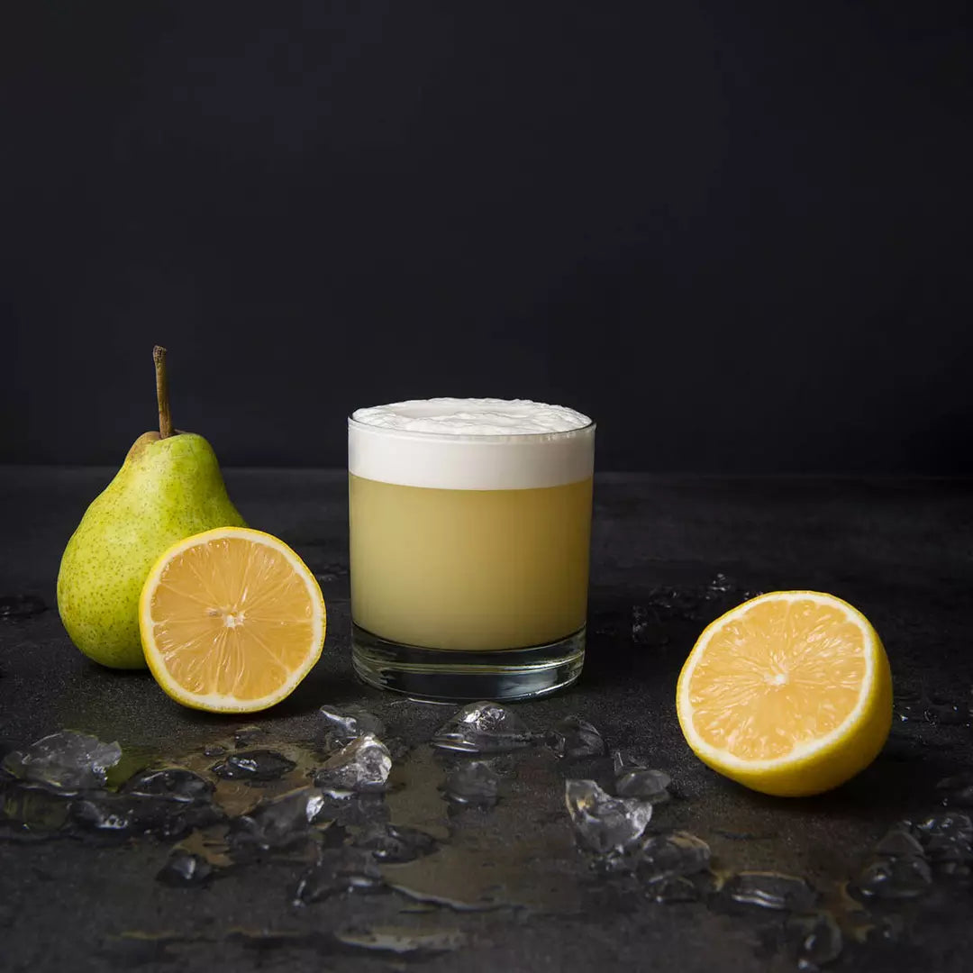 Glass of yellow drink with foam, surrounded by a pear, lemon, and ice on a dark surface.