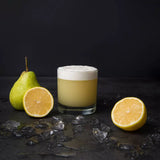 Glass of yellow drink with foam, surrounded by a pear, lemon, and ice on a dark surface.