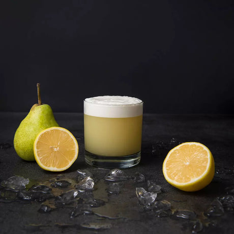 Glass of yellow drink with foam, surrounded by a pear, lemon, and ice on a dark surface.