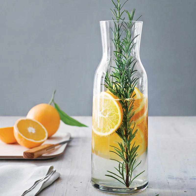 Clear glass carafe with lemon slices and rosemary on a wooden surface with oranges in the background.