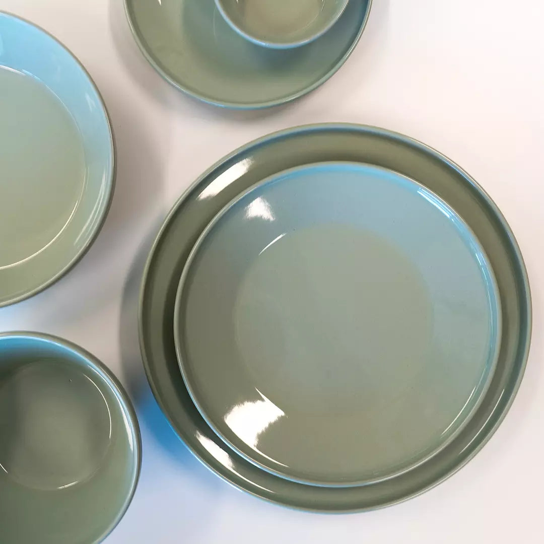 A set of glossy ceramic tableware in a muted sage green color, including a large plate, a smaller plate, a bowl, and a cup, arranged on a white surface.
