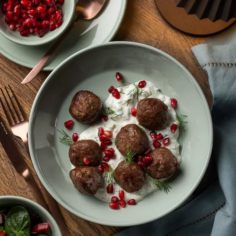 A top-down view of a light green ceramic bowl filled with eight brown meatballs on a white sauce, garnished with pomegranate seeds, dill, and fresh herbs, set on a wooden table with a matching bowl of pomegranates nearby.