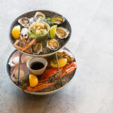 Three-tiered seafood platter with oysters, crab legs, and lemon wedges on a light gray background.