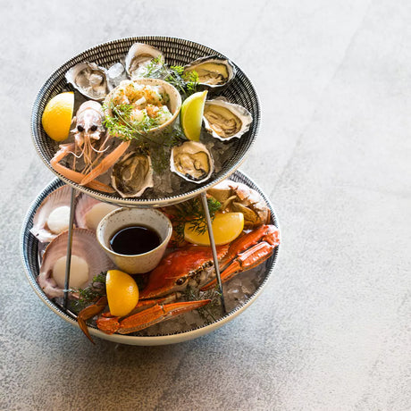 Three-tiered seafood platter with oysters, crab legs, and lemon wedges on a light gray background.