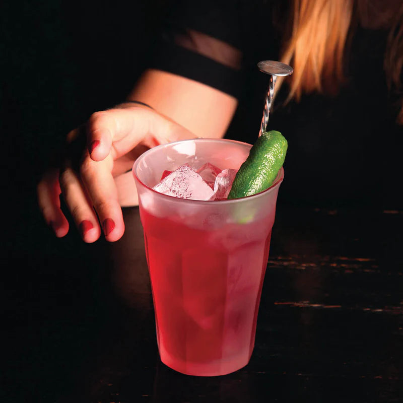 A pink cocktail in a clear plastic cup with ice, garnished with a lime wedge and a metal spoon, held by a person in a dimly lit setting.