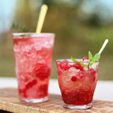 Two glasses of red berry cocktails with ice and garnishes on a wooden surface outdoors.