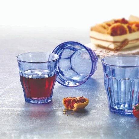 Three blue glasses, one with a red liquid on a metal surface, with waffles in the background.