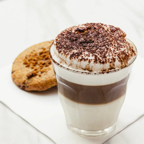 A stunning photo of a creme latte in a small, tapered tumbler with cappuccino top and matching biscuit on a white table setting