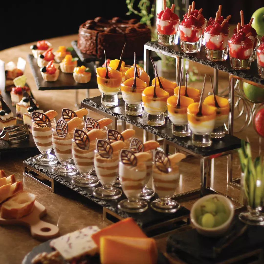 A catering buffet display featuring multiple tiers of dessert cups, fruit, pastries, and cheese on a wooden table in warm ambient lighting.