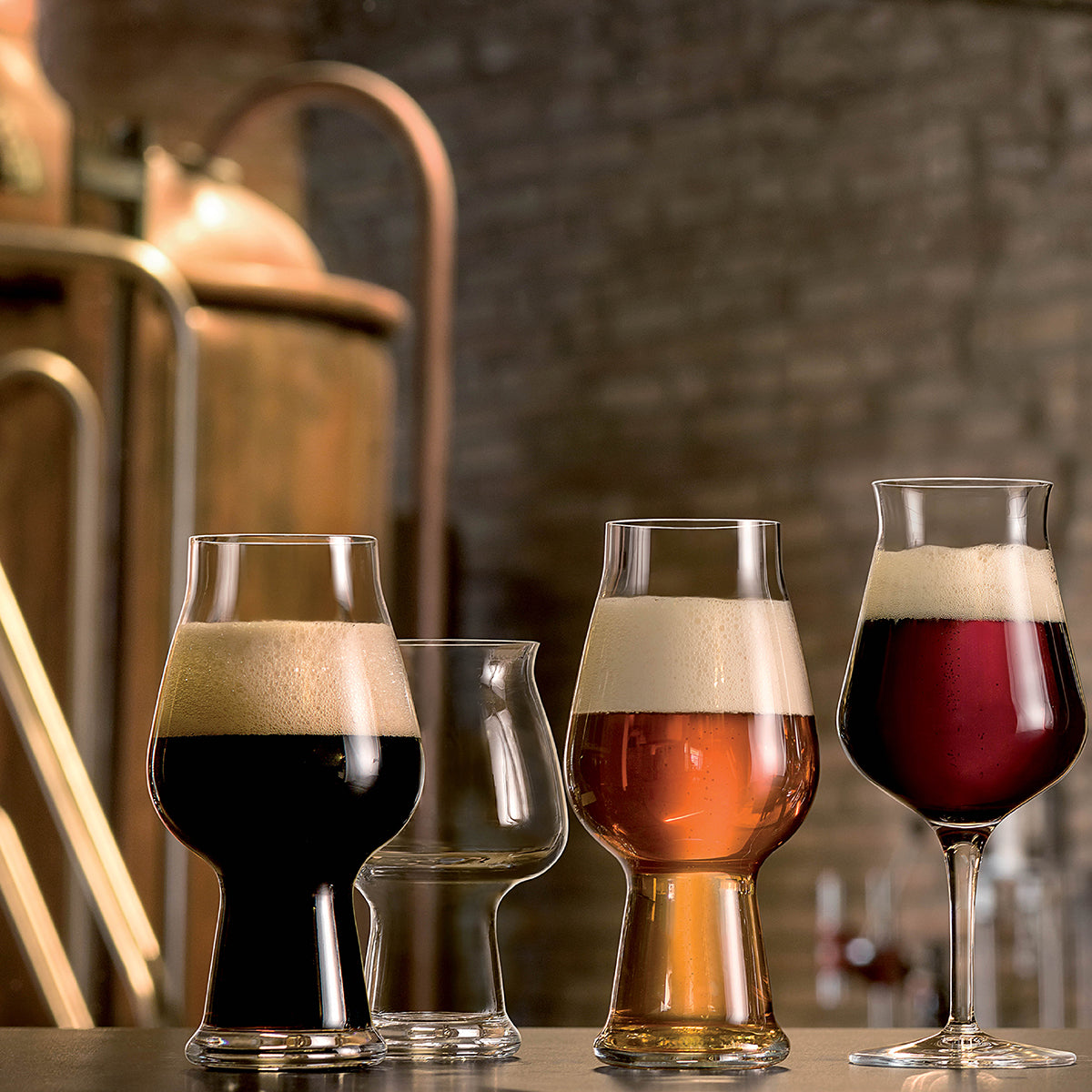 Four different shaped tulip style beer glasses, 3 with liquid and one clear on a table with a brewery background
